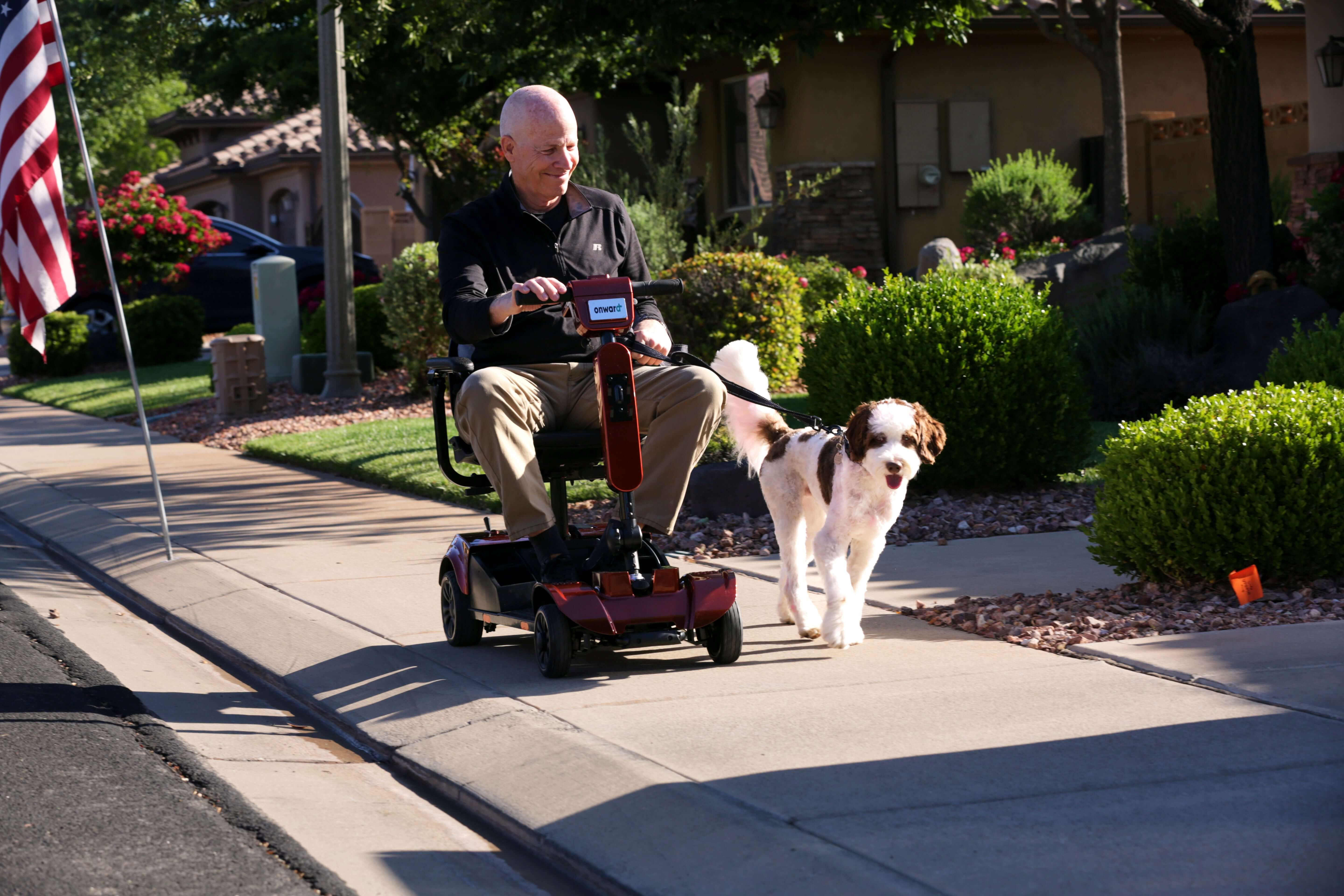 Elderly man enjoying a ride on a mobility scooter while walking his dog along a sunny sidewalk.