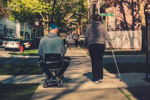 A man on a mobility scooter and a woman with a walking stick stroll on the sidewalk.