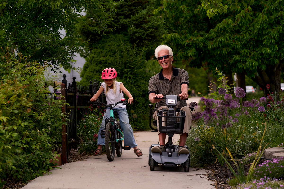 Senior riding an Onward mobility scooter on a garden path beside a child on a bike